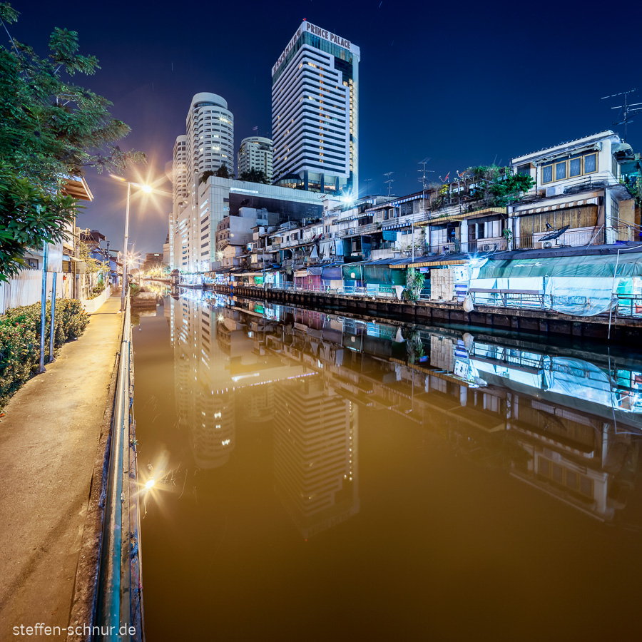 rich and poor
 riverside path
 brown water
 Bangkok
 Thailand
 high rise
 cabins
